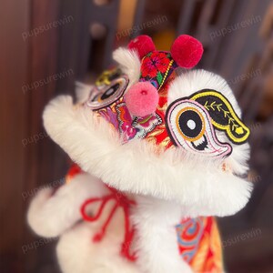 May include: A white plush lion dance costume with red and gold embroidery. The costume has a large, fluffy head with a pink nose and black eyes. The head is decorated with a red and gold embroidered design. The costume is perfect for a Chinese New Year celebration.