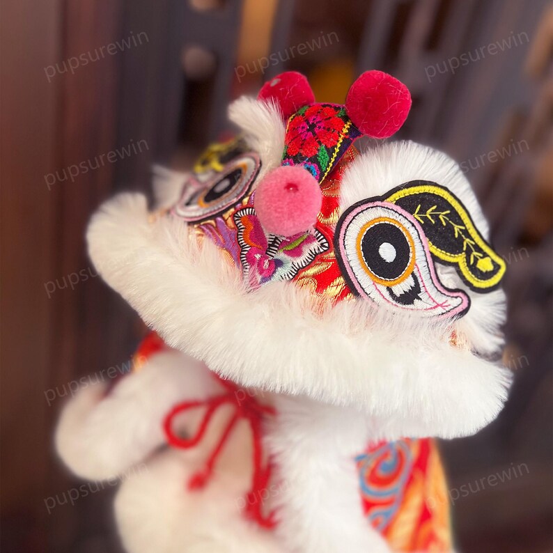 May include: A white plush lion dance costume with red and gold embroidery. The costume has a large, fluffy head with a pink nose and black eyes. The head is decorated with a red and gold embroidered design. The costume is perfect for a Chinese New Year celebration.
