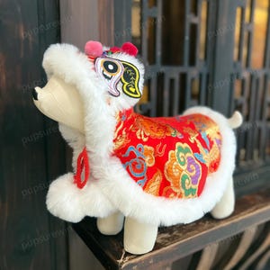 May include: A white and red dog costume with a lion dance head. The costume has a white fur trim and a red fabric with gold and blue embroidery. The lion dance head has a pink pom-pom on top.