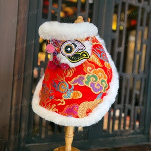 May include: Red and gold Chinese lion dance costume for a pet, with white fur trim and a pink nose.