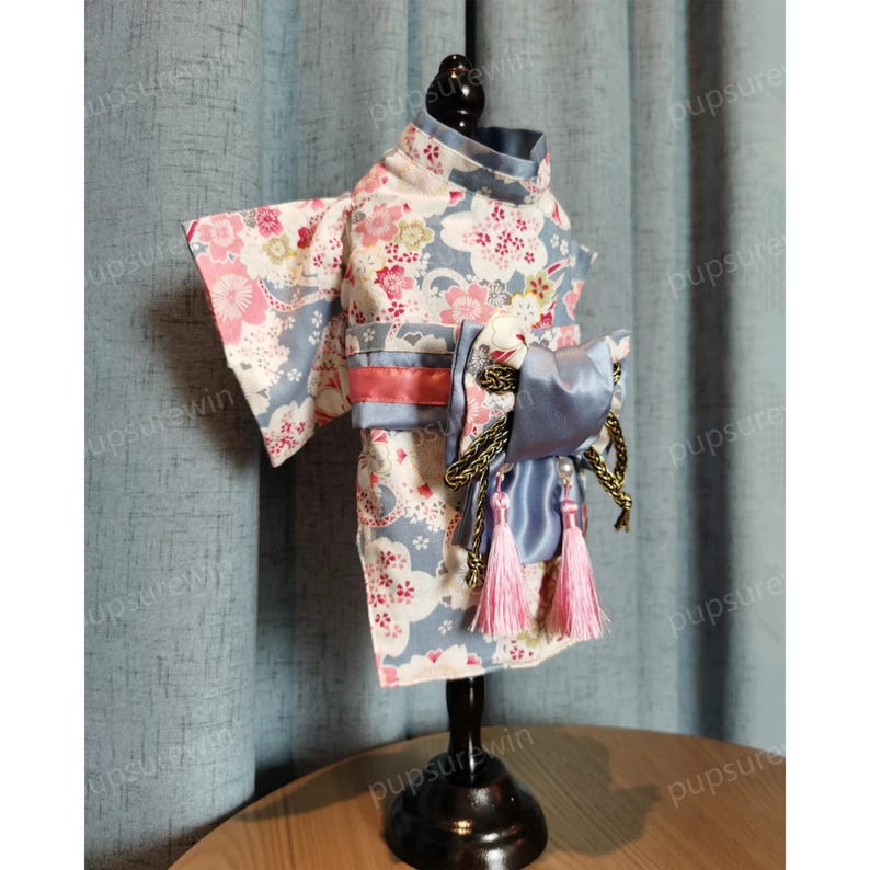 Pu&ograve; includere: Un kimono per cani con motivo floreale blu e bianco con accenti di nastro rosa e blu. Il kimono &egrave; esposto su un supporto nero.