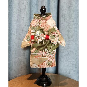 Pu&ograve; includere: Un kimono per cani beige e verde con un fiocco rosso e bianco. Il kimono presenta un motivo floreale e una scena di montagna.