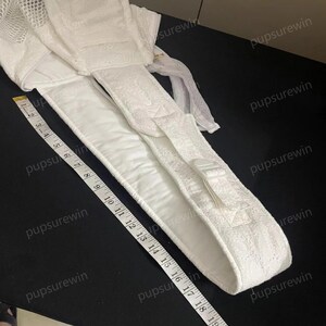 May include: White padded baby carrier strap with a white plastic buckle. The strap is measured with a tape measure that shows 19 inches.