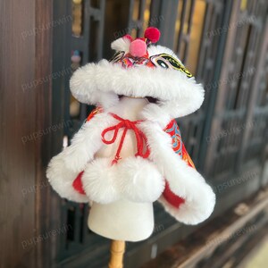 May include: A red and white lion dance costume for a pet. The costume has a white fur hood with a red and gold lion face and a red and gold patterned body. The costume is on a white mannequin.