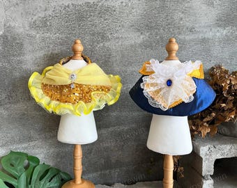 May include: Two pet collars, one yellow with gold sequins and a yellow bow, and one blue with white lace and a blue jewel. Both collars are on white mannequins with wooden stands.
