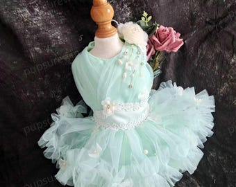 May include: A light green tulle dress for a small dog, featuring a white lace trim, a white satin ribbon, and white flower embellishments. The dress is on a white mannequin and has a white flower on the shoulder.