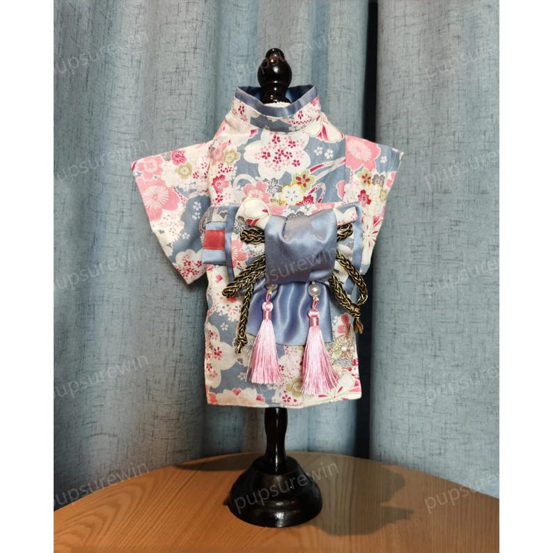 Pu&ograve; includere: Un kimono per cani blu e bianco con stampa floreale rosa e un fiocco di raso blu con bordi dorati e nappe rosa.