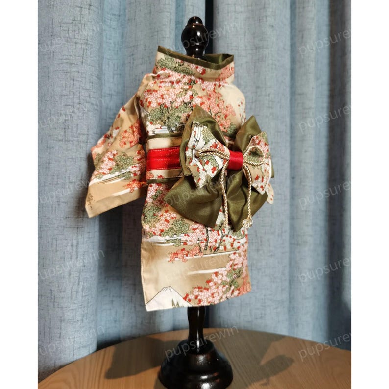 Pu&ograve; includere: Un kimono beige e rosa con un fiocco verde e una cintura rossa per un animale domestico. Il kimono presenta un motivo floreale e una scena di montagna.