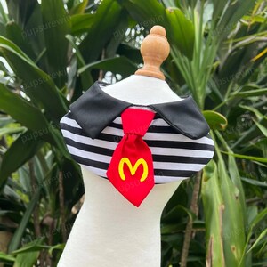 Pode incluir: Um colar de cachorro listrado preto e branco com uma gravata vermelha que apresenta um "M" amarelo no centro. O colar tem um colar preto com uma borda branca.