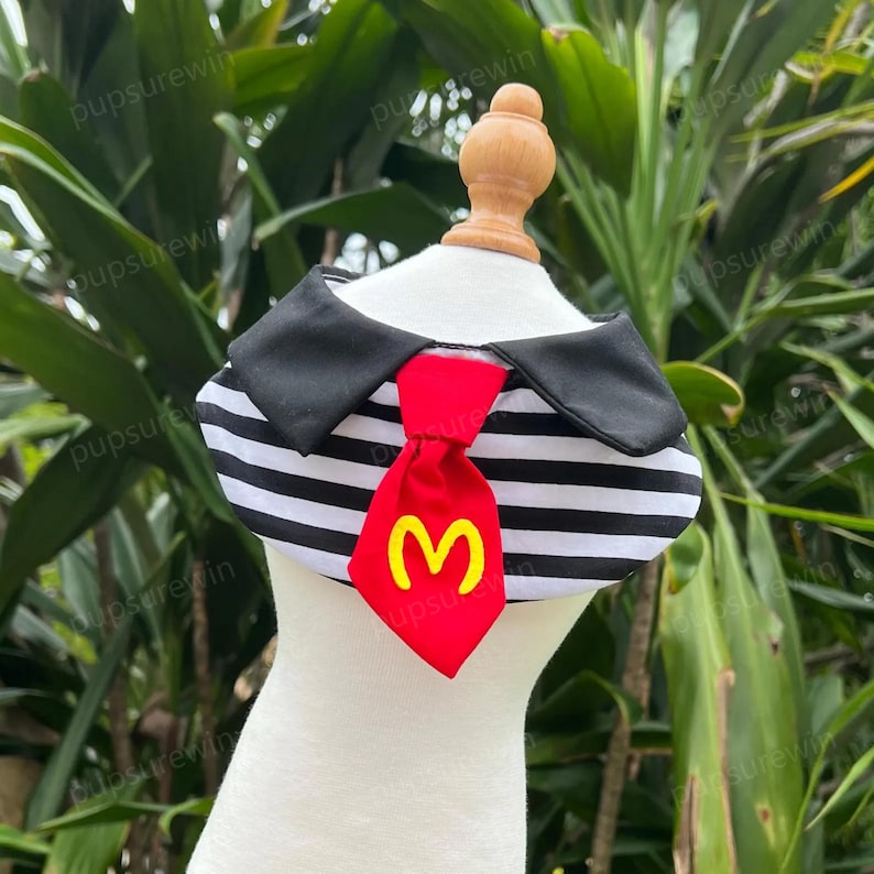 Pode incluir: Um colar de cachorro listrado preto e branco com uma gravata vermelha que apresenta um "M" amarelo no centro. O colar tem um colar preto com uma borda branca.