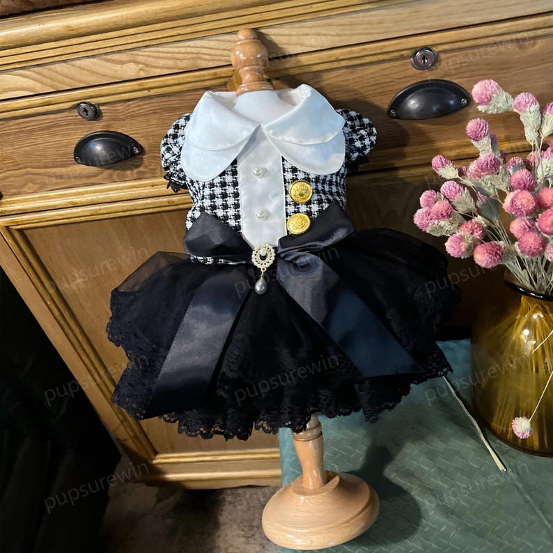 K&ouml;nnte beinhalten: Ein schwarz-wei&szlig;es Hahnentrittkleid mit wei&szlig;em Kragen und schwarzem T&uuml;llrock. Das Kleid ist mit goldenen Kn&ouml;pfen und einer Perlenbrosche verziert. Das Kleid befindet sich auf einer Holzpuppe.