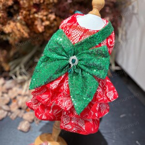 Puede incluir: Vestido de encaje rojo y verde de varias capas con un gran lazo de lentejuelas verdes y un acento de joya verde. El vestido está en un maniquí.