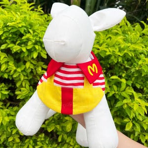 Pode incluir: C&atilde;o de pel&uacute;cia branco vestindo uma camisa listrada vermelha e branca com um babador amarelo e um "M" vermelho no babador.