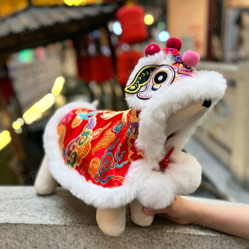 May include: A white and red lion dance costume for a pet. The costume features a white fur hood with a red and gold embroidered lion face and a red satin body with gold embroidery.