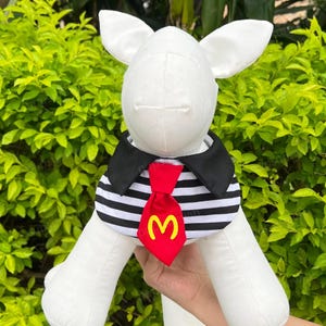 Pode incluir: C&atilde;o de pel&uacute;cia branco vestindo um colar listrado preto e branco com uma gravata vermelha com um logotipo 'M' amarelo.