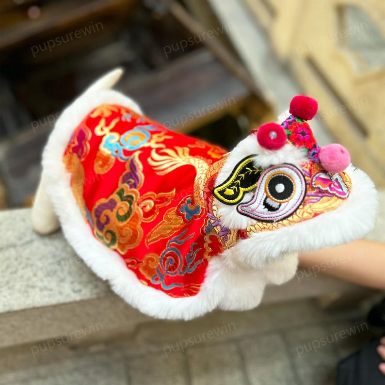 May include: A white dog wearing a red and gold Chinese lion dance costume with white fur trim. The costume has embroidered details and a large, round, black eye.