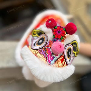 May include: A white and red lion dance head with colorful embroidery and pink pom-poms. The head is made of fabric and fur.