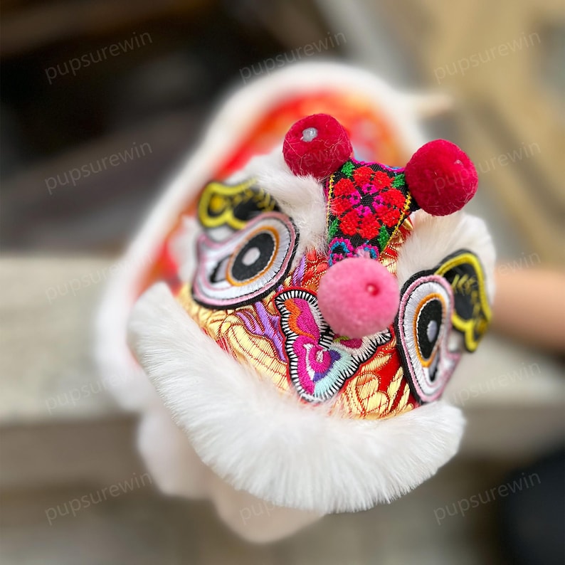 May include: A white and red lion dance head with colorful embroidery and pink pom-poms. The head is made of fabric and fur.