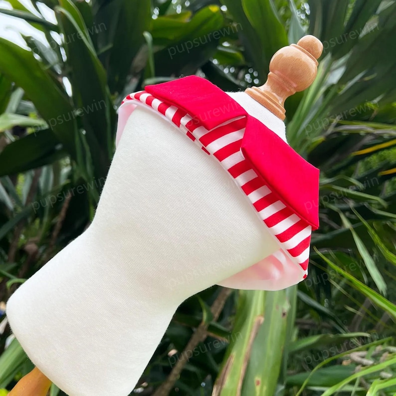 Pode incluir: Um colar branco para animais de estima&ccedil;&atilde;o com um tecido listrado vermelho e branco e um colar de tecido vermelho. O colar est&aacute; em um manequim branco.