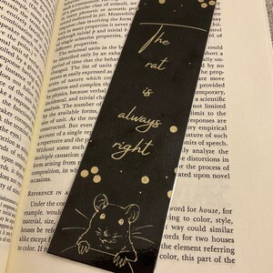 May include: Black bookmark with the text "The rat is always right" in gold lettering. A cartoon rat illustration is at the bottom. The bookmark is resting on an open book.