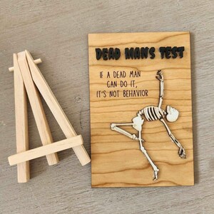 May include: A wooden sign with the text "DEAD MAN'S TEST" and the quote "IF A DEAD MAN CAN DO IT, IT'S NOT BEHAVIOR." A skeleton is depicted in a dancing pose. A small wooden easel is to the left of the sign.