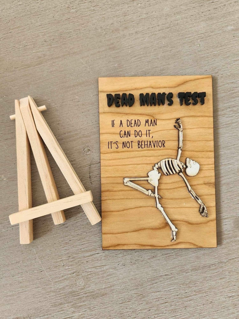 May include: A wooden sign with the text "DEAD MAN'S TEST" and the quote "IF A DEAD MAN CAN DO IT, IT'S NOT BEHAVIOR." A skeleton is depicted in a dancing pose. A small wooden easel is to the left of the sign.