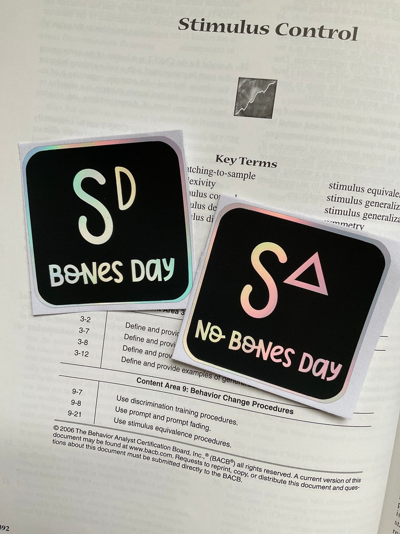 May include: Two black square stickers with iridescent edges. One reads "SD BONES DAY" in white script, the other reads "SA NO BONES DAY". The stickers are on a textbook page with the title "Stimulus Control".