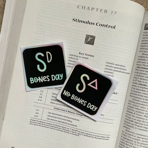 May include: Two square stickers with iridescent edges are placed on an open textbook. One sticker reads "SD BONES DAY" in a playful font, and the other says "SA NO BONES DAY". The stickers are black with white and light green text.