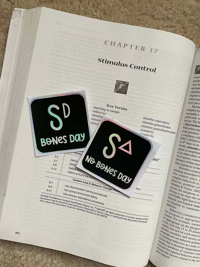 May include: Two square stickers with iridescent edges are placed on an open textbook. One sticker reads "SD BONES DAY" in a playful font, and the other says "SA NO BONES DAY". The stickers are black with white and light green text.