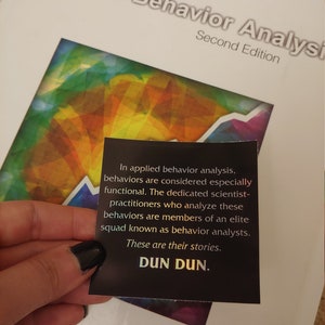 May include: A close-up of a black sticker with white text that reads "In applied behavior analysis, behaviors are considered especially functional. The dedicated scientist-practitioners who analyze these behaviors are members of an elite squad known as behavior analysts. These are their stories. DUN DUN."