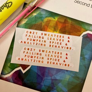 May include: Two white stickers with orange text that reads "Cozy sweaters & falling leaves, pumpkin spice & analyzing behavior." The stickers are on a colorful background.