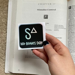May include: A square sticker with a black background featuring the letters "SA" and a triangle in white, with the words "No Bones Day" in white below. The sticker is placed on an open book.