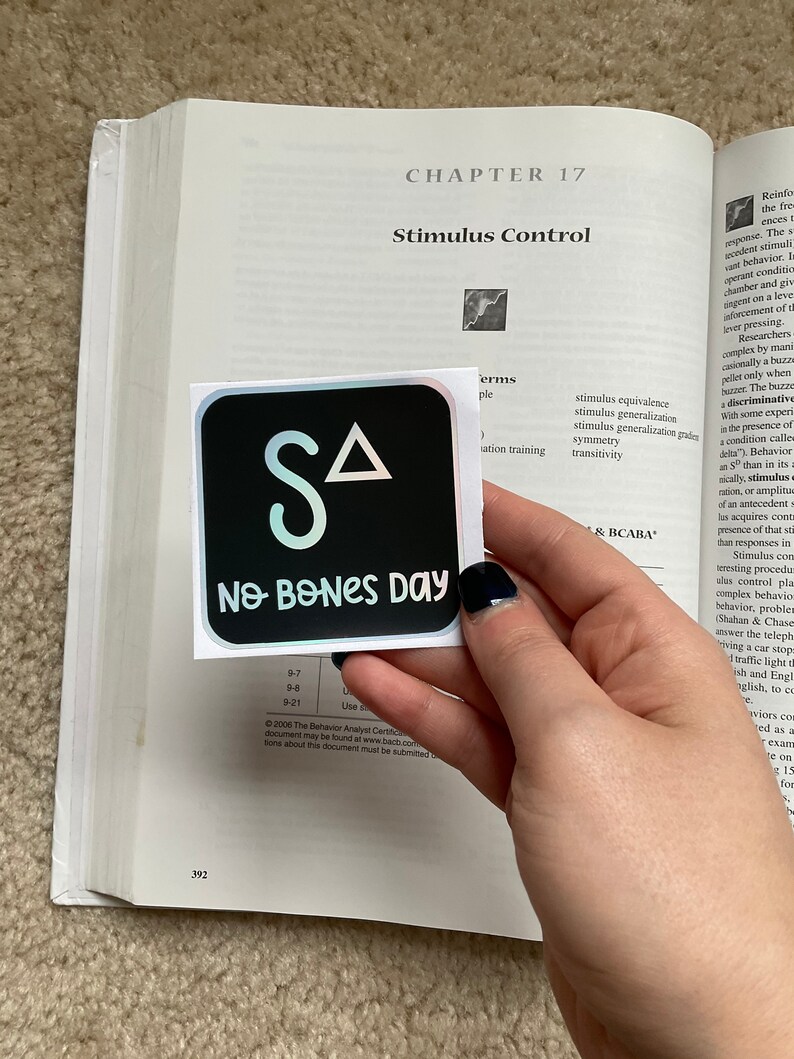 May include: A square sticker with a black background featuring the letters "SA" and a triangle in white, with the words "No Bones Day" in white below. The sticker is placed on an open book.