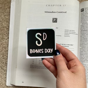 May include: A black square sticker with the text "SD BONES DAY" in a whimsical font. The sticker is held over an open textbook with the title "Stimulus Control" visible. The background is a beige carpet.