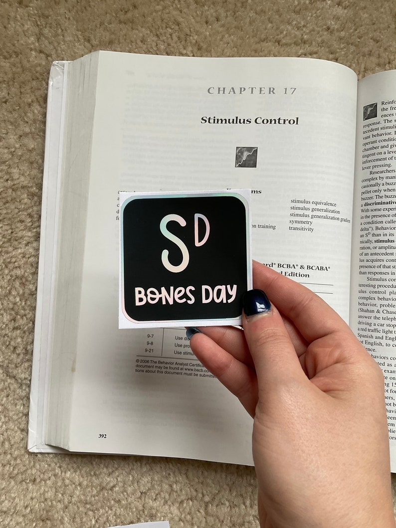 May include: A black square sticker with the text "SD BONES DAY" in a whimsical font. The sticker is held over an open textbook with the title "Stimulus Control" visible. The background is a beige carpet.