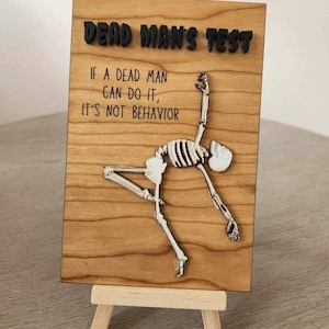 May include: A wooden sign with the text "DEAD MAN'S TEST" and the quote "IF A DEAD MAN CAN DO IT, IT'S NOT BEHAVIOR." A skeleton figure is depicted in a dynamic pose. The sign is displayed on a small wooden easel.