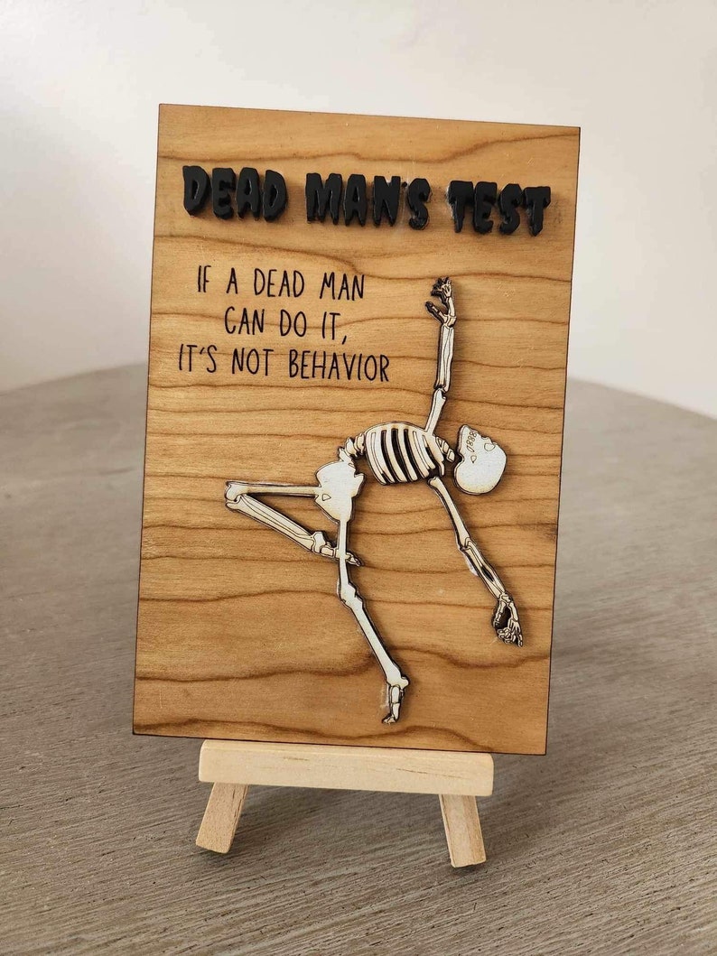 May include: A wooden sign with the text "DEAD MAN'S TEST" and the quote "IF A DEAD MAN CAN DO IT, IT'S NOT BEHAVIOR." A skeleton figure is depicted in a dynamic pose. The sign is displayed on a small wooden easel.
