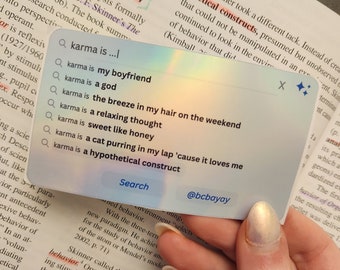 May include: A close-up of a hand holding a smartphone with a search bar displaying the text "karma is..." and a list of auto-suggested search terms. The search bar is highlighted in blue and the suggested terms are in black text. The background is a page of text from a book about behavior.