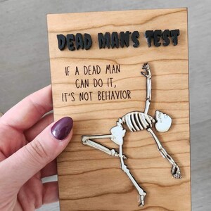 May include: Wooden plaque with the text "DEAD MAN'S TEST" and the quote "IF A DEAD MAN CAN DO IT, IT'S NOT BEHAVIOR." A skeleton figure is depicted in a yoga pose. The text and skeleton are in black and white.