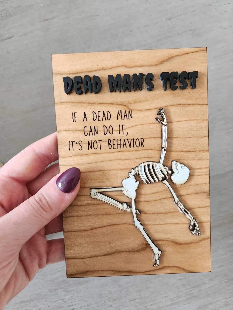 May include: Wooden plaque with the text "DEAD MAN'S TEST" and the quote "IF A DEAD MAN CAN DO IT, IT'S NOT BEHAVIOR." A skeleton figure is depicted in a yoga pose. The text and skeleton are in black and white.