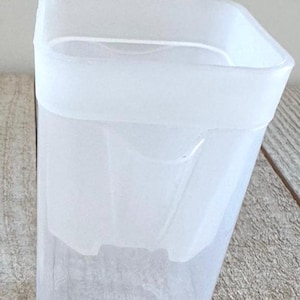 May include: A translucent, frosted plastic container with a square shape and rounded corners. The vessel features a distinct inner rim structure and a smooth, matte finish, presenting a simple, functional design for storage or organization purposes.