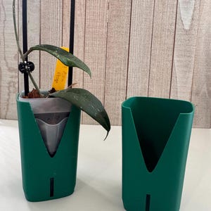 May include: Two dark green plastic plant pots with a V-shaped cutout on the front and a vertical drainage slot at the base. One pot contains a plant with dark green leaves, a black metal arch trellis, and a yellow plant identification tag.