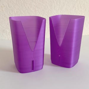 May include: Two identical purple plastic holders with a semi-translucent finish. Each holder features a wide V-shaped cutout at the top rim and a small vertical rectangular slot positioned near the base on the front side of the object.