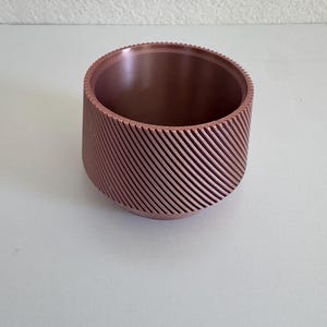 May include: A cylindrical bowl with a metallic copper finish and a distinct diagonal ribbed texture along the exterior walls. The interior is smooth and features a consistent color throughout, with a flat base and a circular opening at the top.