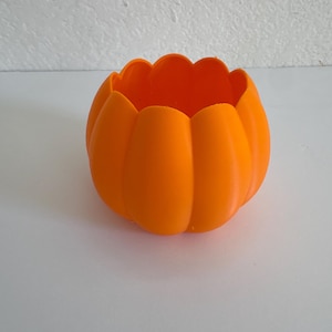 May include: A bright orange, hollow, pumpkin-shaped container with a smooth, matte finish. The object features a rounded, bulbous body with vertical ridges and a scalloped, open top edge, resembling the natural form of a small, round pumpkin.
