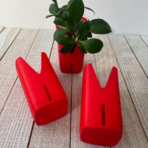 May include: Three bright red, tooth-shaped planters with a smooth finish. Each planter features a vertical rectangular slot on the front and a deep V-shaped opening at the top. One planter contains a small green plant with thick, dark leaves.