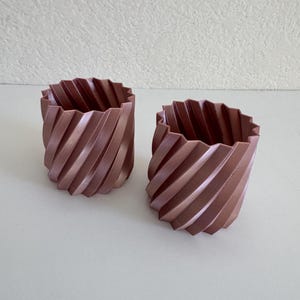 May include: Two cylindrical containers with a metallic rose gold finish and a distinct spiral, pleated texture along the exterior walls. Each vessel features an open top with a jagged, star-like rim, creating a uniform, repeating geometric pattern.