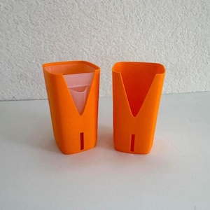 May include: Two orange plastic cups with a V-shaped cutout on the front and a vertical slit near the base. One cup contains a translucent light pink inner liner, while the other cup is empty, showing the bright orange interior and exterior.