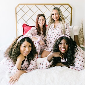 Può includere: Quattro donne indossano pigiami rosa con stampa di Babbo Natale e posano su un letto bianco. Sorridono tutte e guardano la fotocamera. I pigiami hanno maniche lunghe e una chiusura con bottoni sul davanti. Le donne indossano fasce per capelli con un fiocco.