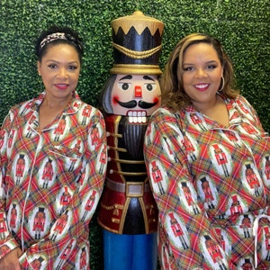 May include: Two people wearing red and white plaid pajamas with a nutcracker print. The pajamas have a button-up front and long sleeves. The people are standing in front of a green wall with a large nutcracker doll.